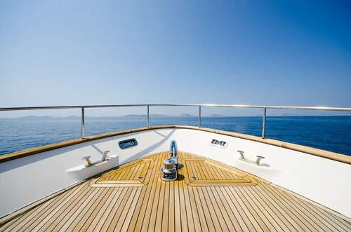 A wooden deck of a boat with a beautiful view of blue waters, distant mountains, and a cloudless blue sky.